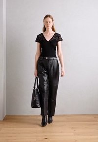 Body noir avec dentelle, pantalon en cuir taille haute, et un sac à main noir. Le mannequin porte des bottines noires à talons aiguilles. Sol en bois.