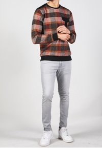 Uomo che indossa un maglione a quadri arancione, marrone e nero, jeans slim-fit grigio chiaro e sneakers bianche, in piedi con le mani incrociate.