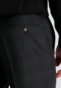 Close-up van een donkere, geruite pantalon met een zak en knoop, gedragen met een zwart topje, waarbij een deel van de hand aan de rechterkant zichtbaar is.