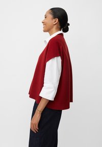 Gilet rosso lavorato a maglia con maniche a cappuccio, realizzato in tessuto morbido, indossato sopra una camicia bianca con bottoni, abbinato a jeans scuri. Design semplice e moderno.