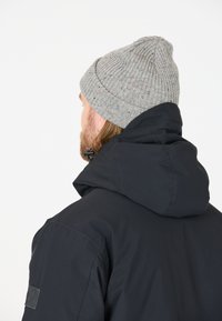 Schwarze isolierte Jacke mit Kapuze, kombiniert mit einer hellgrauen Strickmütze mit bunten Sprenkeln, die Textur und Wärme ausstrahlt.
