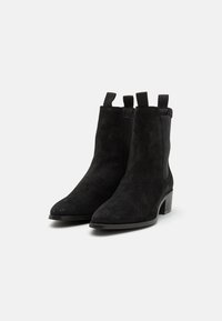 Botas de ante negras hasta el tobillo con punta afilada, paneles laterales elásticos y tiradores. El tacón es de forma cuadrada y de altura moderadamente baja.