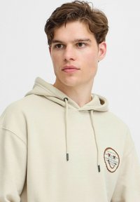 Felpa con cappuccio in cotone beige, con tasca frontale e cappuccio regolabile con cordino. Presenta un patch ricamato rotondo con testo e dettagli a filo in contrasto.