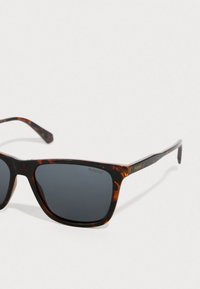 Γυαλιά ηλίου καστανής απόχρωσης με σκούρους φακούς και ορθογώνιο σχήμα. Διαθέτουν μεταλλικές λεπτομέρειες και σήμα "Polaroid" στους βραχίονες.