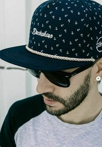Gorra marina con patrón de ancla blanca, con el logotipo bordado "Blackskies" y un detalle de cuerda. Llevada con gafas de sol negras, combinada con una camiseta.