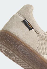 Gros plan sur le talon d'une chaussure beige Adidas avec une étiquette en tissu Cordura et une semelle extérieure en caoutchouc brun texturée, estampillée du logo Adidas.