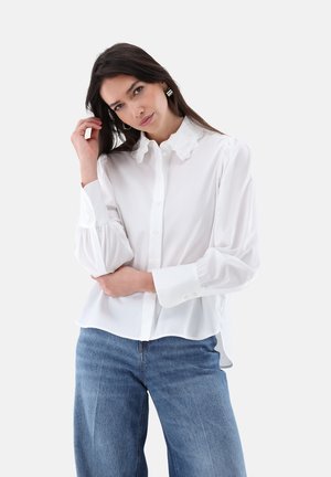 Donna dai capelli scuri che indossa una camicetta bianca con bottoni e colletto in pizzo e jeans blu, che posa con una mano che le accarezza i capelli.