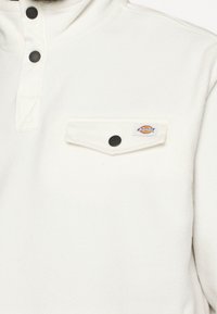 Biały polarowy sweter z kołnierzykiem zapinanym na pół guzika, z kieszenią na piersi oraz czarnymi guzikami, o miękkiej fakturze i subtelnym logotypie na kieszeni.