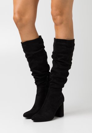 Botas largas de ante negras con un diseño holgado, punta redonda y tacón grueso; cuentan con un cremallera lateral para facilitar su uso.