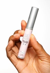 Läppolja i ett ljusrosa rör med ett silverlock, med djärv svart text. Hållen i handen mot en vit bakgrund.