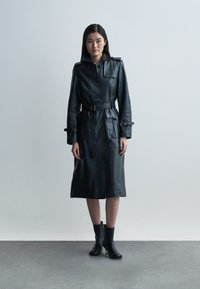 Black leather trench coat with a belted waist, button closure, and side pockets. Features a wide collar and structured silhouette.