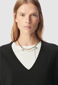 Kobieta z prostymi blond włosami nosi warstwowe złote i srebrne łańcuchy na białej koszuli i czarnym swetrze w serek na jasnym tle.