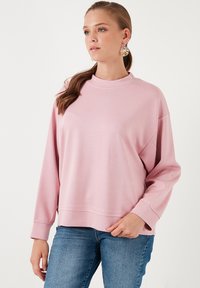 Felpa rosa con collo alto, spalle cadenti e maniche lunghe. Tessuto liscio, vestibilità rilassata. Abbinata a jeans blu.