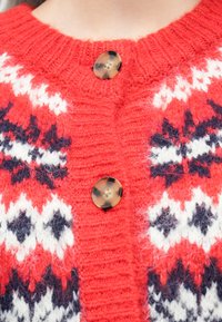 Κόκκινη πλεκτή ζακέτα με γεωμετρικό μοτίβο σε ναυτικό μπλε και λευκό, με στρογγυλό κουμπί από χελώνα στο λαιμό. Υφή υφάσματος.