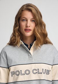 Szaro-kremowa koszulka polo w paski z haftem "POLO CLUB" w kolorze granatowym. Posiada kołnierzyk z kontrastowym kolorem wewnętrznym i miękką teksturę tkaniny.