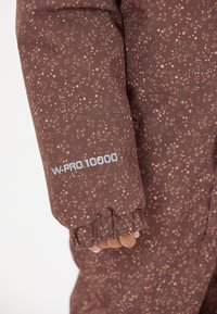 Brun jakke med blomsterdesign og tekstureret finish, med elastiske manchetter og et "W-PRO 10000" mærke i lyseblå på ærmet.