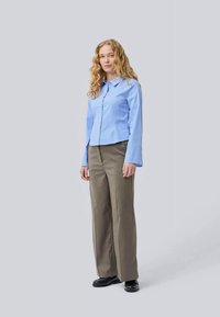 Camicia button-up a righe verticali blu chiaro, colletto, abbinata a pantaloni beige a gamba larga e scarpe nere. Modello in piedi contro uno sfondo grigio.
