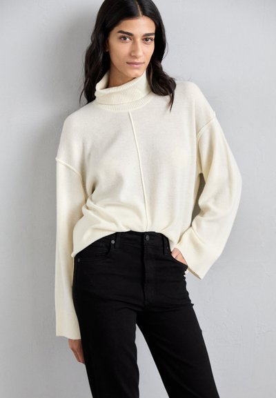 Kremowy, oversize'owy sweter z wysokim kołnierzem, miękką teksturą i prążkowanymi mankietami, w połączeniu z dopasowanymi czarnymi dżinsami. Prosty design, bez wzorów.
