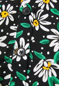 Tissu noir avec imprimé de fleurs de marguerite blanches, feuilles vertes, petits points blancs et boutons noirs cousus sur une patte de boutonnage.