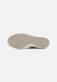 Semelle de sneaker blanche avec un caoutchouc beige texturé arborant un logo en relief. Le design inclut deux rainures horizontales pour la flexibilité.