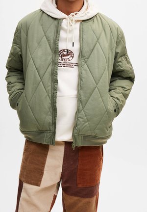 Personne portant une veste matelassée vert clair sur un sweat à capuche blanc avec texte et un pantalon en velours côtelé patchwork marron, crème et rouille.