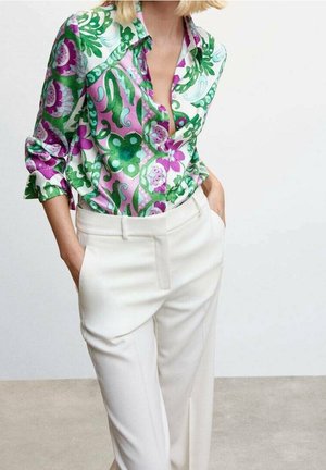 Femme portant un pantalon blanc taille haute, les mains dans les poches, et une chemise à manches longues avec des motifs floraux verts et violets audacieux.