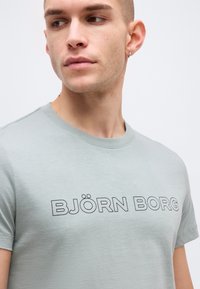 Νεαρός άνδρας με κοντά μαλλιά και σκουλαρίκι που φοράει ανοιχτό γκρι μπλουζάκι με το κείμενο "BJÖRN BORG" τυπωμένο στο στήθος, κοιτάζοντας προς τα πλάγια.