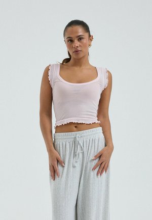 Nő hátrafogott hajjal, világos rózsaszín, rövidített ujjú toppal és bő, világosszürke, megkötős nadrággal, egyszerű háttér előtt állva.