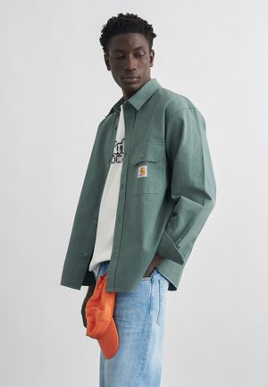 Joven con camisa verde abotonada sobre una camiseta blanca estampada y vaqueros azul claro, sosteniendo una gorra naranja a su lado.