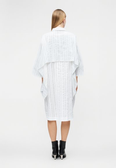 Γυναίκα που στέκεται με την πλάτη στην κάμερα, φορώντας λευκό φόρεμα μέχρι το γόνατο με διαφανείς κάθετες ρίγες και λεπτομέρεια με βολάν καπέλου, μαύρα μποτάκια αστραγάλου.