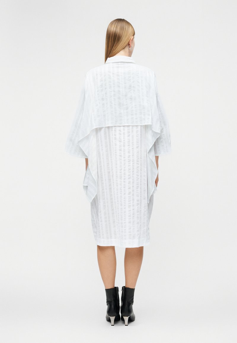 Femme vue de dos portant une robe blanche mi-longue avec des rayures verticales transparentes et un détail cape à volants, bottines noires.