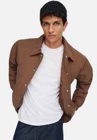 Braune gesteppte Jacke mit Kragen, silberne Druckknöpfe, über einem weißen T-Shirt getragen, kombiniert mit dunklen Denim-Jeans. Schwarze Strickmütze auf dem Modell.