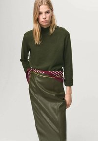 Maglione lavorato a maglia verde oliva con scollatura a coste e dettagli con bottoni. Abbinato a una gonna di pelle e una cintura bordeaux a righe.