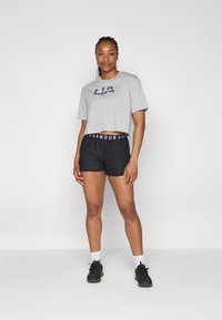 Camiseta gris corta con logo "UA", combinada con pantalones cortos deportivos negros con una cinturilla de logo. El modelo lleva zapatillas negras y calcetines blancos.