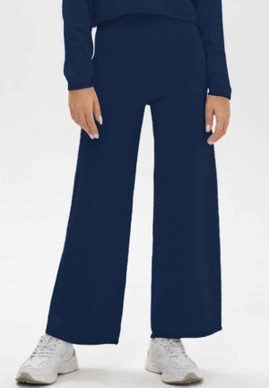 Persona che indossa pantaloni larghi blu navy, top a maniche lunghe abbinato e sneakers bianchi, in piedi su uno sfondo neutro.