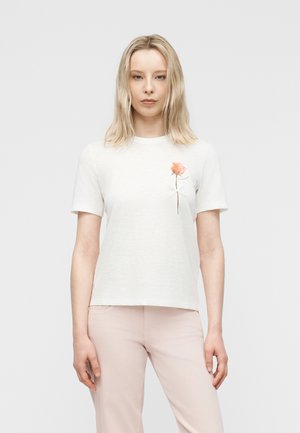 Femme blonde portant un t-shirt blanc avec un motif de fleur orange sur le côté gauche de la poitrine, associée à un pantalon rose clair, se tenant devant un fond blanc.