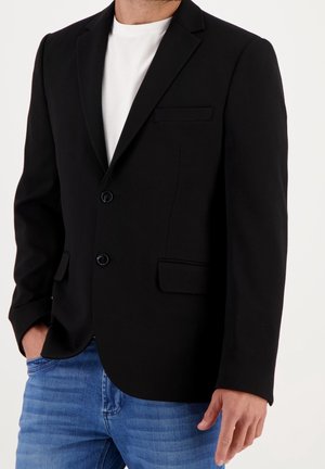 Man draagt een zwarte blazer over een wit t-shirt met blauwe jeans, een hand in de zak, tegen een effen lichte achtergrond.
