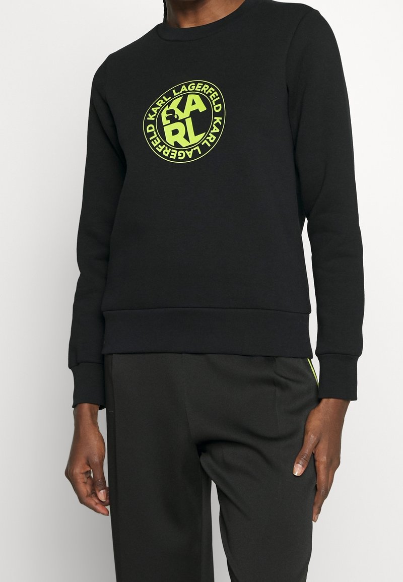 Personne portant un sweat-shirt noir avec un logo circulaire Karl Lagerfeld jaune néon sur la poitrine et un pantalon noir assorti.