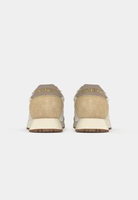 Beige og creme sneakers med en ruskindsbag, guldbogstaver på hælen og en textured gummisål. Klassisk lavprofil design.
