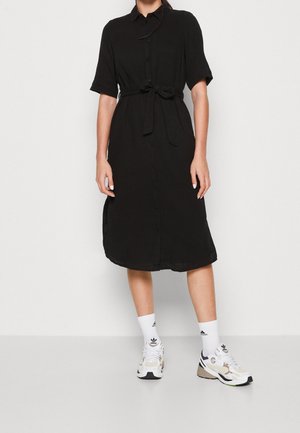 Femme portant une robe noire boutonnée mi-longue avec manches courtes, ceinture nouée à la taille, chaussettes blanches Adidas et baskets blanches.