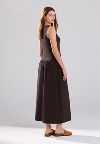 Robe longue en coton brun foncé avec un haut ajusté et une jupe froncée. Le modèle porte des sabots marron clair. Texture lisse, design sans manches, accents simples.