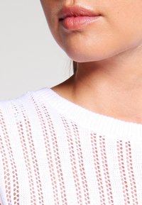 Pull blanc en maille avec un design en maille ajourée, présentant des rayures horizontales et verticales. Texture douce et encolure large. Gros plan sur la zone du col.