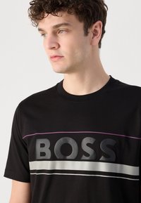 Fekete póló kerek nyakkivágással, nagy "BOSS" logóval szürke és fehér csíkokkal, a logó felett lila vonallal kiemelve.