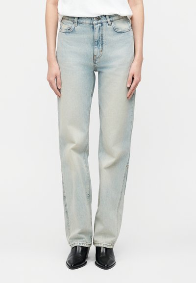 Personne portant un jean droit bleu clair à taille haute, associé à des bottes noires à bout pointu et un haut blanc rentré.
