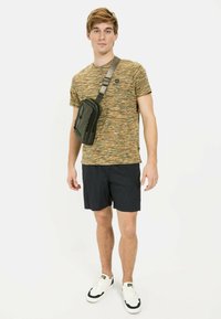 Zielona nylonowa torba na ramię z zamkiem, noszona na kolorowej, w paski koszulce z krótkim rękawem i granatowych krótkich spodniach, w zestawie z białymi sneakersami.