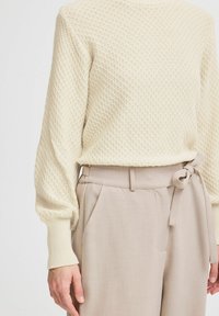 Pull en tricot beige clair avec un motif texturé, poignets côtelés, porté par-dessus un pantalon beige pâle à ceinture nouée.