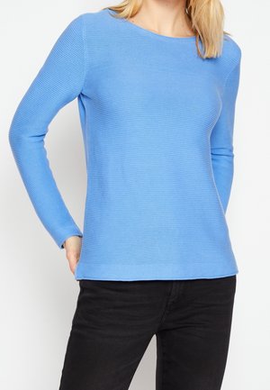 Femme portant un pull côtelé bleu clair à manches longues avec un pantalon noir, mains posées sur les hanches, vue du cou aux hanches.