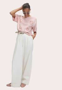 Roze korte mouw t-shirt met getextureerde bloemaccenten, gecombineerd met wijde, witte broek en lichtroze instapschoenen.