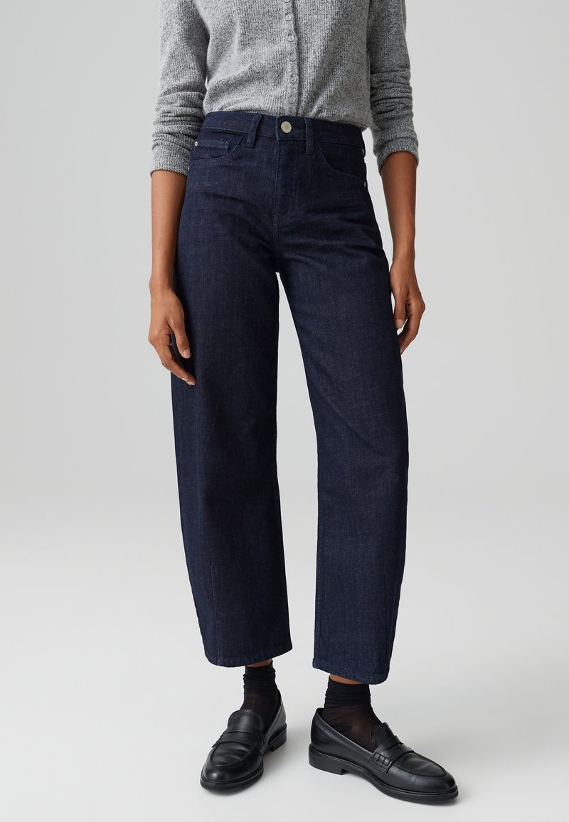Personne portant un jean bleu foncé à jambes larges, un haut gris à manches longues boutonné, des chaussettes noires et des mocassins en cuir noirs, debout devant un fond uni.