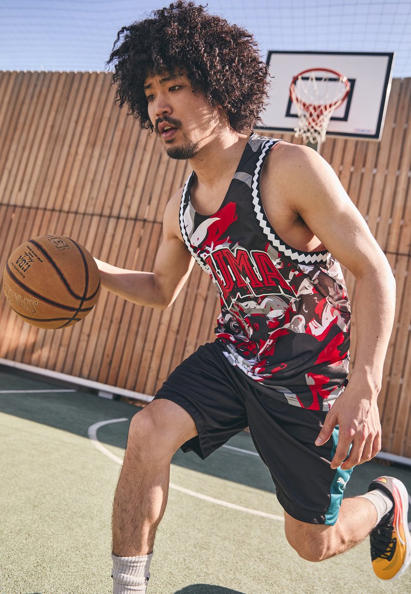 Basketbalista v živě barevném, vzorovaném tílku a černých šortkách, dribluje hnědý basketbal na hřišti s dřevěným pozadím.